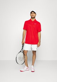 Rød polo t-shirt med krave og Nike-logo, parret med hvide shorts, holder en tennisketsjer, iført hvide og lyserøde sneakers.