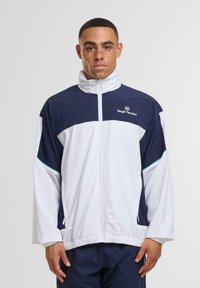 Rövid hajú és szeplős férfi, aki sötétkék és fehér sportdzsekit visel, ami fel van zárva, és egy sima fehér háttér előtt áll.