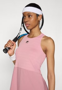 Pink tennis dress with a textured upper, smooth lower section, racerback design, and Fila logo; completed with a white headband and wristband.