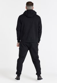 Černá mikina s velkou kapucí, žebrovanými manžetami a odpovídajícími kapsáčovými kalhotami s bočními kapsami, doplněná černými botami, pohled zezadu.