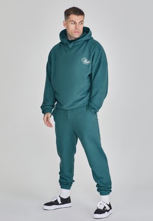 Teal hoodie with front pocket and logo, paired with matching tapered joggers. Model wears black and white sneakers with white crew socks.