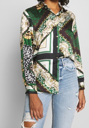 Femme portant une chemise boutonnée à manches longues à motifs verts, noirs, blancs et dorés, associée à un jean bleu clair déchiré.