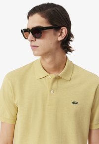 Geel polo shirt van een gestructureerde stof met een drieknoopsluiting, voorzien van een groen geborduurd logo. Zwarte zonnebril met getinte lenzen.