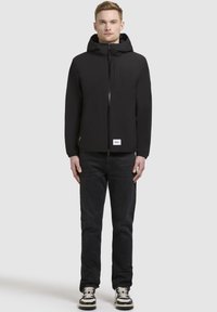 Black hooded jacket with a front zipper, featuring a chest pocket and elastic cuffs, paired with dark jeans and black-and-white sneakers.