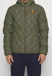 Mand iført en olivengrøn quiltet zip-up jakke med hætte og ellesse-logoer på brystet, parret med navy blå bukser.