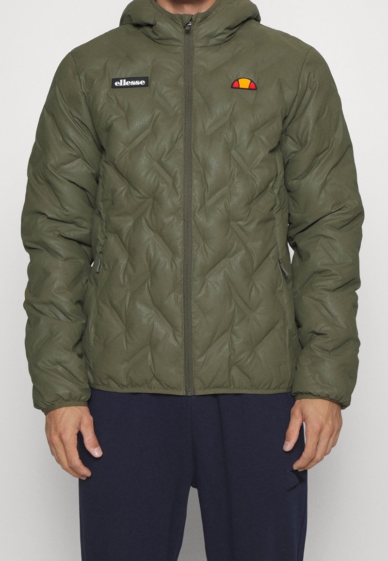 Mand iført en olivengrøn quiltet zip-up jakke med hætte og ellesse-logoer på brystet, parret med navy blå bukser.