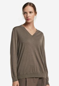 Kvinna med ljus hy och brunt hår bär en brun V-ringad långärmad tröja och matchande bruna byxor, och ser något åt sidan.