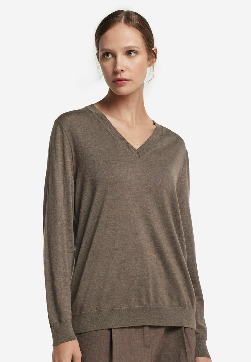 Kvinna med ljus hy och brunt hår bär en brun V-ringad långärmad tröja och matchande bruna byxor, och ser något åt sidan.