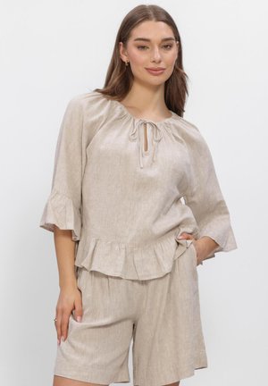 Femme portant un chemisier en lin beige avec des manches à volants et un ourlet, assorti à un short ample, posant avec une main dans la poche.