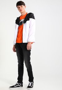 Coupe-vent léger bicolore en blanc et noir, associé à un t-shirt orange, un jean noir et des baskets noires.