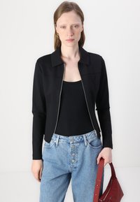 Black zippered jacket with a collared neckline and long sleeves, paired with a black tank top and light blue high-waisted jeans.
