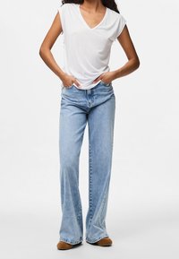 Kvinna iklädd en vit V-ringad t-shirt, ljusblå vida jeans och bruna skor, stående med händerna i fickorna framför en slät vit bakgrund.