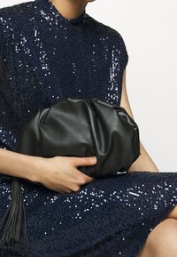 Black leather clutch bag with a gathered design and tassel detail, held in a hand. Background features a navy sequined dress.