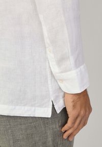 Camicia di lino bianca con polsini arrotolati, caratterizzata da un dettaglio con bottoni. Tessuto con trama visibile, abbinata a pantaloni grigi.
