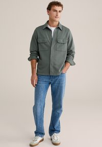 Zöld, gombos ing, puha anyagból készült, két mellzsebbel, világoskék farmernadrággal és fehér sportcipővel párosítva.