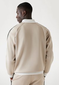 Beige sportjack met een ribbelige witte kraag, zwarte zijkanten en een gladde textuur. Heeft raglanmouwen en een aansluitende taille.