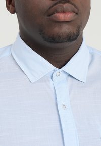 Jasnoniebieska koszula zapinana na guziki, wykonana z teksturowanej tkaniny, z klasycznym kołnierzykiem i srebrnymi guzikami. Widok z bliska na kołnierzyk i listwę z guzikami.