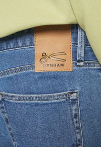 Jean en denim bleu avec un patch en cuir marron au-dessus de la poche arrière montrant un logo de ciseaux et le nom de la marque Denham.