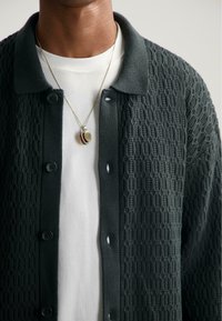 Donkergroene gebreide cardigan met een textuurpatroon, met een knopenlijst aan de voorkant en een kraaghalslijn, gedragen over een wit shirt.
