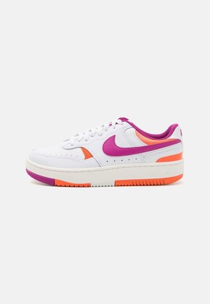 Baskets basses blanches avec des accents rose et orange, un logo Nike violet, des lacets blancs, et une semelle blanche épaisse avec une bande de roulement orange et violette.