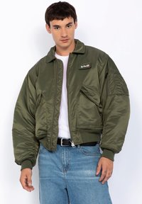 Olive green bomber jacket with a zip front, ribbed cuffs, and collar. Features a logo patch on the chest and zippered pocket on the sleeve.
