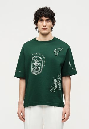 Jeune homme aux cheveux bouclés portant un t-shirt oversize vert foncé avec des imprimés blancs de fontaines et de fleurs et un pantalon blanc.