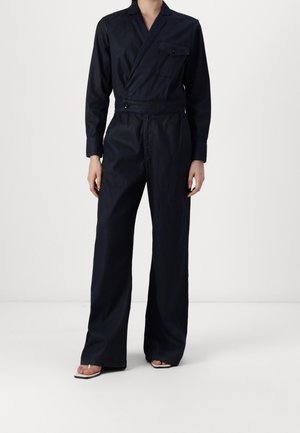 Person trägt einen dunkelmarineblauen Wickel-Jumpsuit mit langen Ärmeln, weitem Bein, einer vorderen Tasche und offenen Sandaletten mit Absatz.