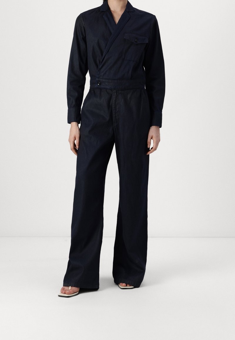 Personne portant une combinaison style cache-cœur bleu marine foncé, manches longues, jambes larges, poche avant et sandales à talons ouvertes.