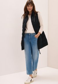 Schwarze Daunenweste mit einem Frontreißverschluss, hohem Kragen und seitlichen Taschen, getragen über einem hellen Pullover und kombiniert mit weiten blauen Jeans und weißen Sneakers.