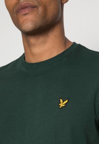 Grünes Baumwoll-T-Shirt mit Rundhalsausschnitt und einem kleinen gelben gestickten Vogel-Logo auf der linken Brustseite. Glatte Textur.