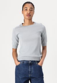Pulover scurt cu mânecă scurtă, de culoare gri deschis, cu decolteu rotund, cu manșete și tiv țesute, fabricat dintr-un material moale, purtat cu blugi cu talie înaltă.