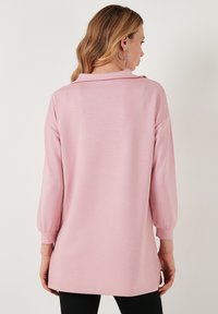 Maglietta lunga a maniche rosa chiaro con colletto, caratterizzata da una vestibilità comoda, fessure laterali e polsini a coste. Tessuto morbido, design casual.