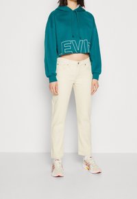 Hoodie court bleu sarcelle avec imprimé blanc "LEVI'S", associé à un jean droit écru et des baskets claires avec des accents roses.