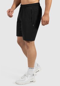 Zwarte sportshorts met een rekbare tailleband, twee zijzakken met rits en een lengte boven de knie, gecombineerd met witte sneakers en sokken.