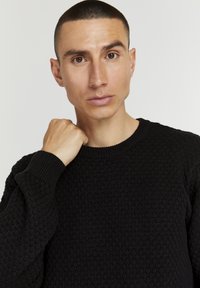 Svart strikkegenser med et teksturert mønster og ribbede mansjetter. Rund hals og lange ermer gir en tettsittende look.