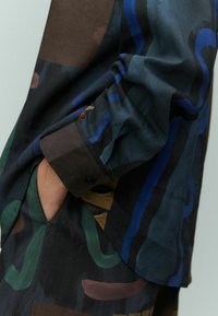Camicia multicolore con motivi astratti in tonalità scure di verde, blu e marrone. Il tessuto sembra morbido e presenta un dettaglio con polsino abbottonato.