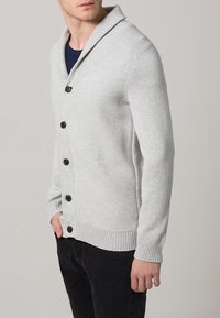 Šedý pletený cardigan s šálovým límcem, s knoflíkovou přední částí, žebrovanými manžetami a texturovaným vzorem. Kombinován s černými kalhotami.