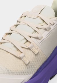 Κοντινή λήψη αθλητικού παπουτσιού κρεμ με υφαντό ύφασμα για το επάνω μέρος, κορδόνια κρεμ και μωβ σόλα.