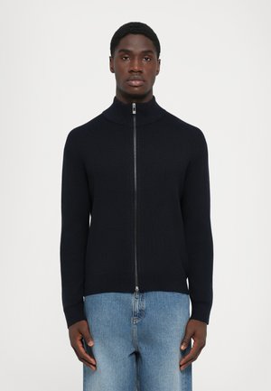 Mann iført en svart ribbestrikket jakke med glidelås og ståkrage, og lyseblå jeans, stående mot en ensfarget hvit bakgrunn.