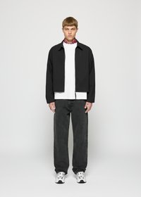 Ung mann står mot hvit bakgrunn iført svart jakke, hvit t-skjorte, mørke jeans, hvite sneakers og rød mønstret skjerf.