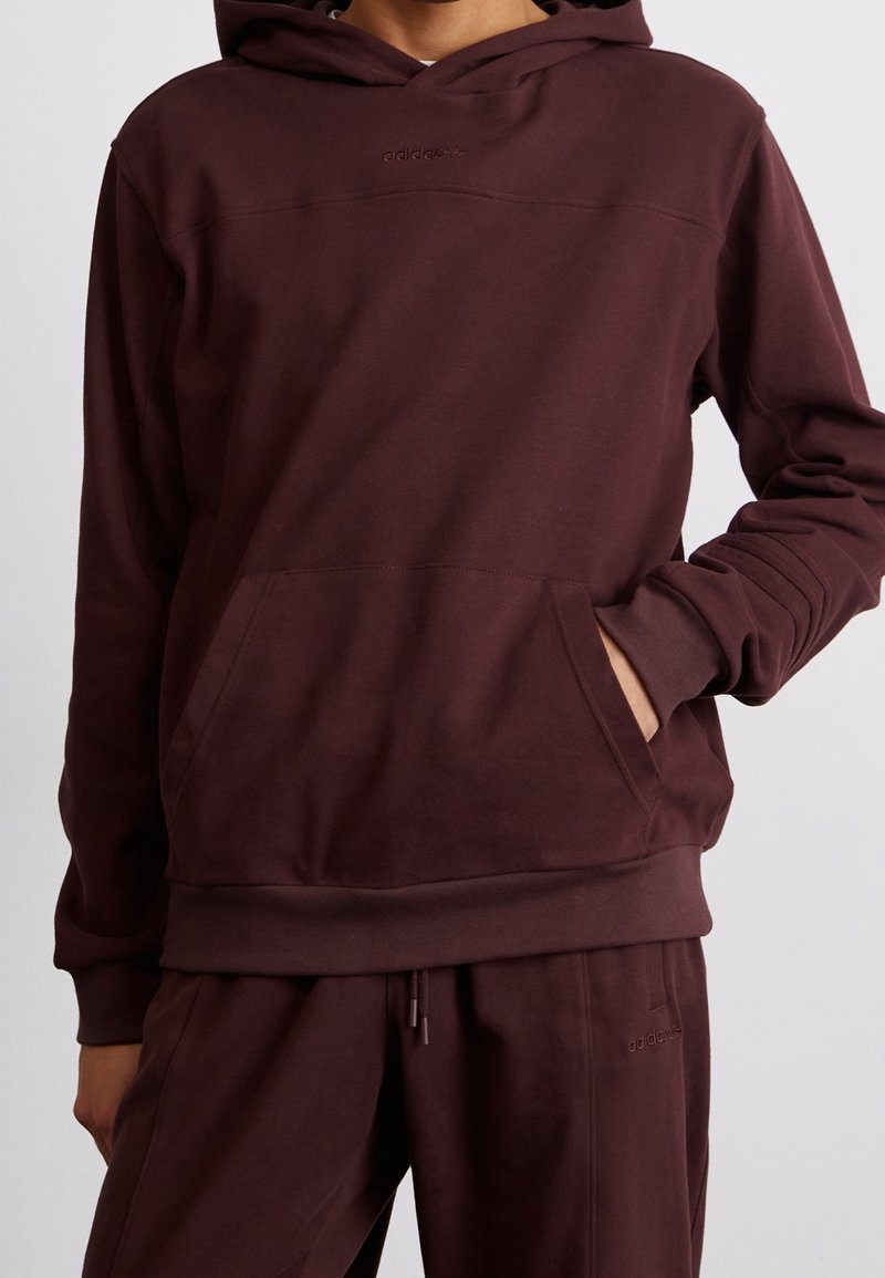 Burgundinvärinen huppari, jossa on etutasku, ribbettikäsivarsit ja helma. Kuoressa on hienovarainen logo yksityiskohtana rinnassa ja sileä kangas rakenne.