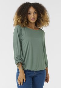 Green, long-sleeve blouse with a round neckline and gathered detail at the cuffs, made of soft material, paired with blue jeans.