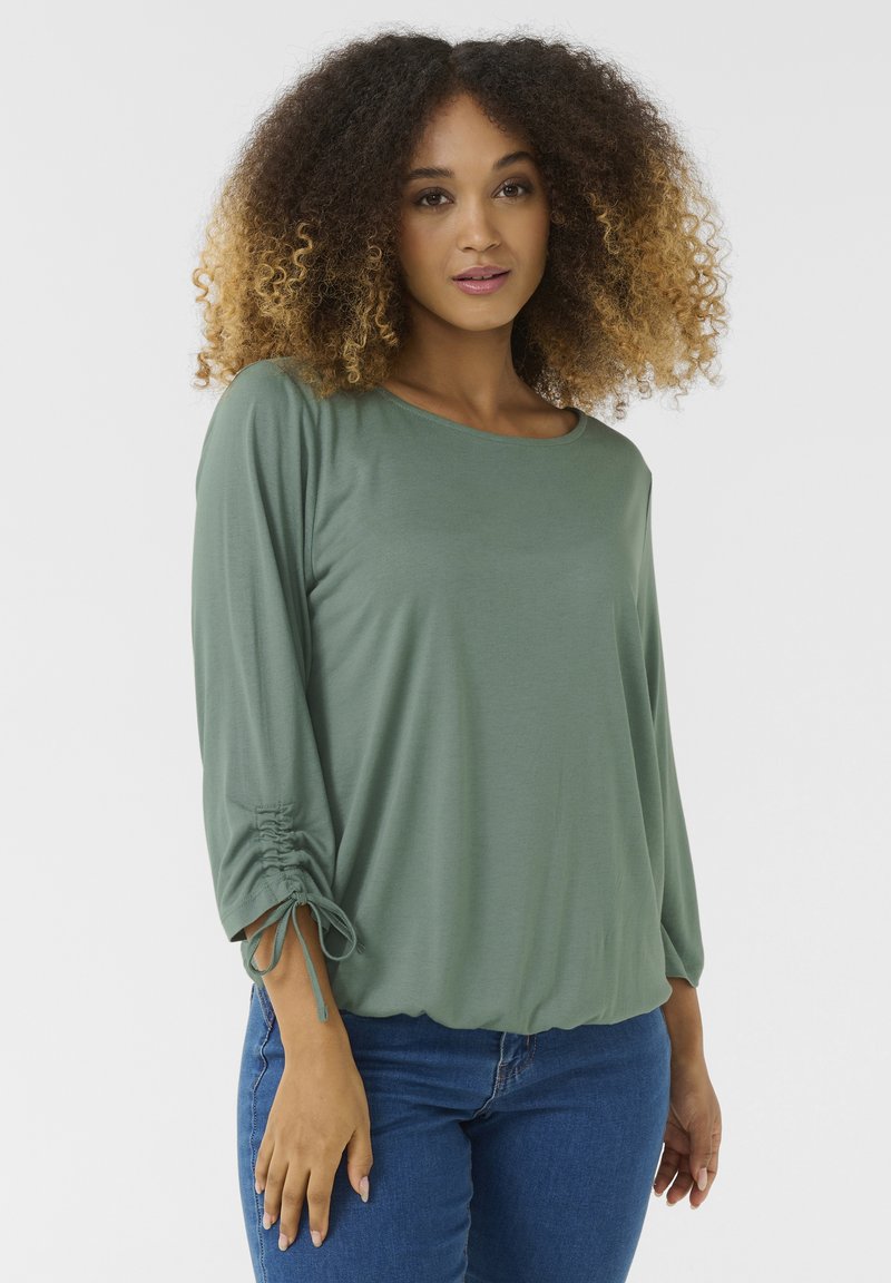 Green, long-sleeve blouse with a round neckline and gathered detail at the cuffs, made of soft material, paired with blue jeans.