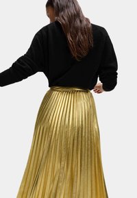 Sort sweater med lange ærmer over en metalisk gylden, plisseret nederdel. Nederdelen har en skinnende overflade og en tætsiddende talje.