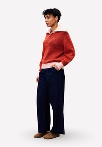 Pull en laine orange et rose à fermeture éclair quart de zip, pantalon en denim foncé à jambes larges, et chaussons en cuir brun texturé, avec des poignets et un col côtelés.