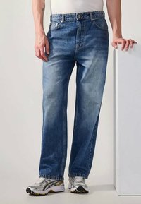 Ανοιχτό μπλε τζιν παντελόνι με χαλαρή εφαρμογή, με ξεθωριασμένη υφή, ίσια πόδια, τρύπες ζωνών και φθορές, συνδυασμένο με γκρι και μαύρα αθλητικά παπούτσια.