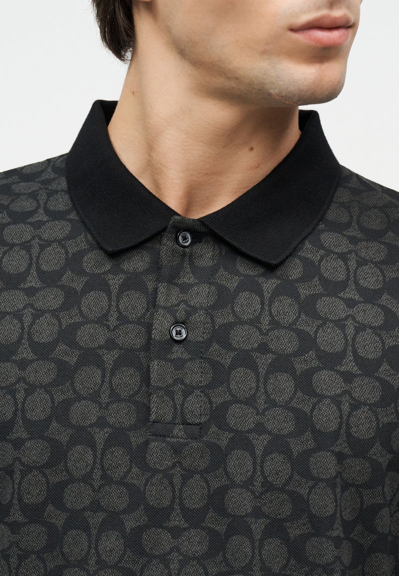 Homme portant un polo noir à motifs avec deux boutons noirs et un col noir uni, regardant sur le côté.