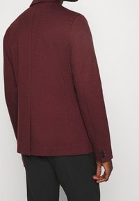 Bordowa marynarka o teksturowanej tkaninie, z klapą z wcięciem, mankietami na jeden guzik i dopasowanym krojem. Brak widocznych wzorów czy zdobień.