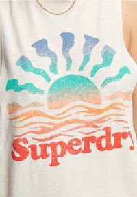 Biała koszulka bez rękawów z grafiką zachodu słońca w odcieniach teal, pomarańczowym i niebieskim, z napisem "Superdry" w pogrubionej czerwieni u dołu.
