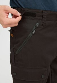 Schwarze Cargo-Hosen mit einem Klettverschlussbund, einer seitlichen Reißverschlusstasche und Knopf-Details. Der Stoff wirkt strapazierfähig und hat eine glatte Textur.